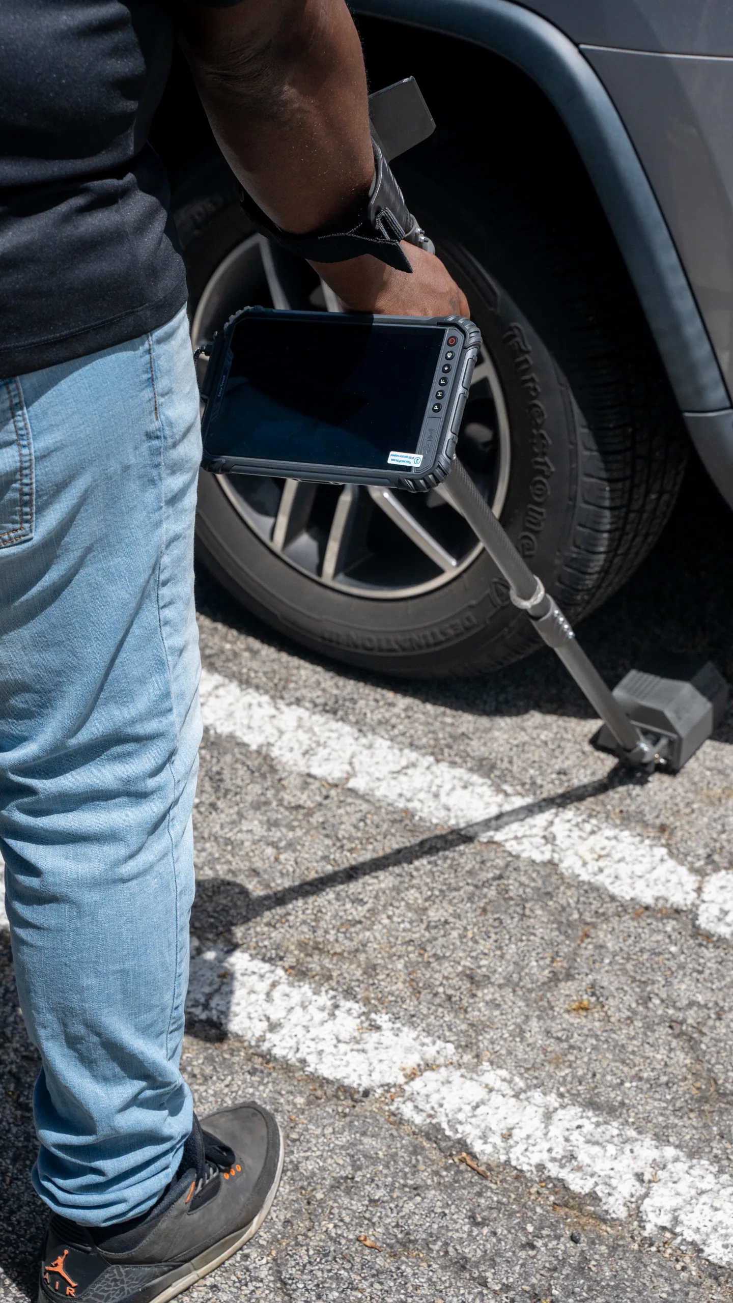 Person using a device with a long pole to inspect a car tire in a parking lot.